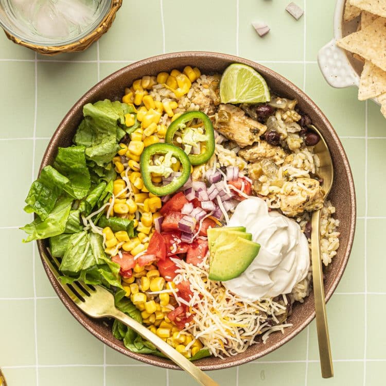 A bowl with a gold fork and spoon and white rice, chicken, black beans, cheese, corn, lettuce, tomatoes, diced onions, sour cream, lime wedges, and sliced jalapeno.