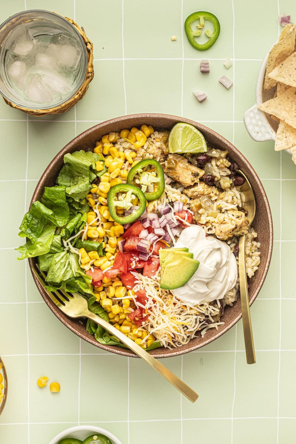 A bowl with a gold fork and spoon and white rice, chicken, black beans, cheese, corn, lettuce, tomatoes, diced onions, sour cream, lime wedges, and sliced jalapeno.