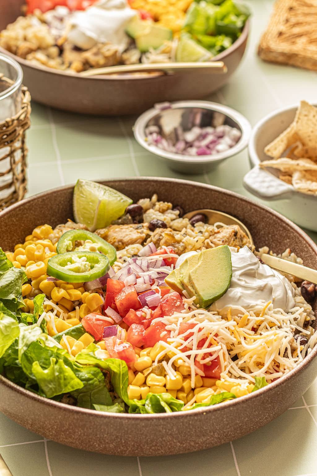 Close up image of a bowl with chicken, rice, sour cream, shredded cheese, lettuce, tomatoes, avocado, sliced jalapeno, and lime wedges.