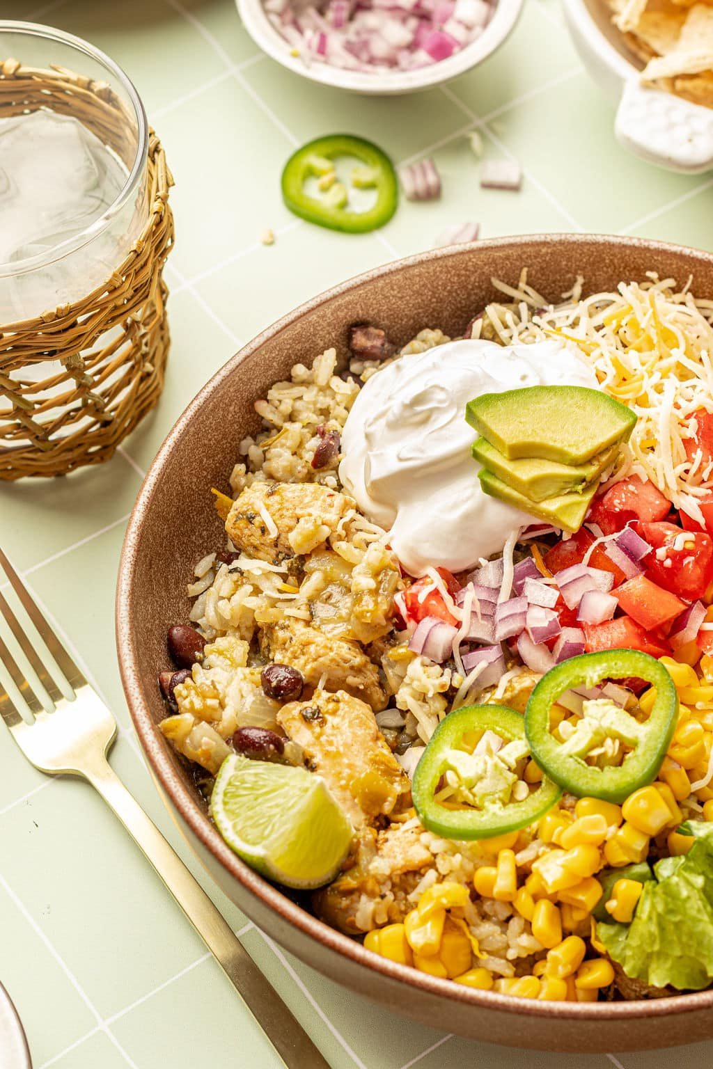 Close up image of a bowl with chicken, rice, sour cream, shredded cheese, lettuce, tomatoes, avocado, sliced jalapeno, and lime wedges.