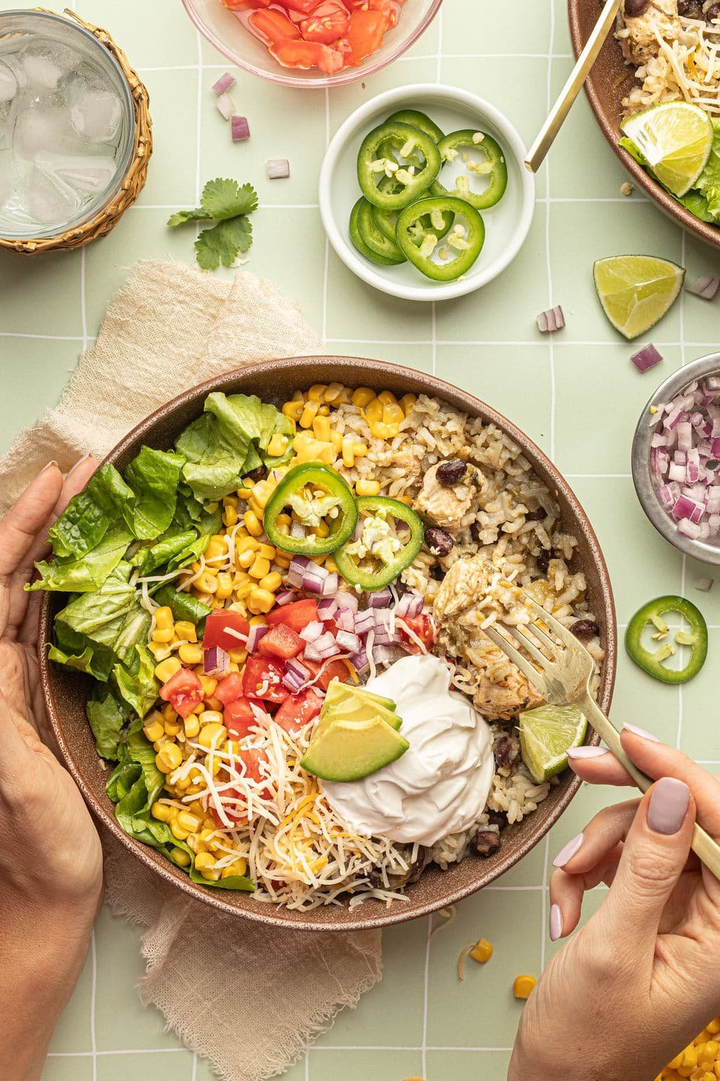 A bowl with a gold fork and spoon and white rice, chicken, black beans, cheese, corn, lettuce, tomatoes, diced onions, sour cream, lime wedges, and sliced jalapeno.