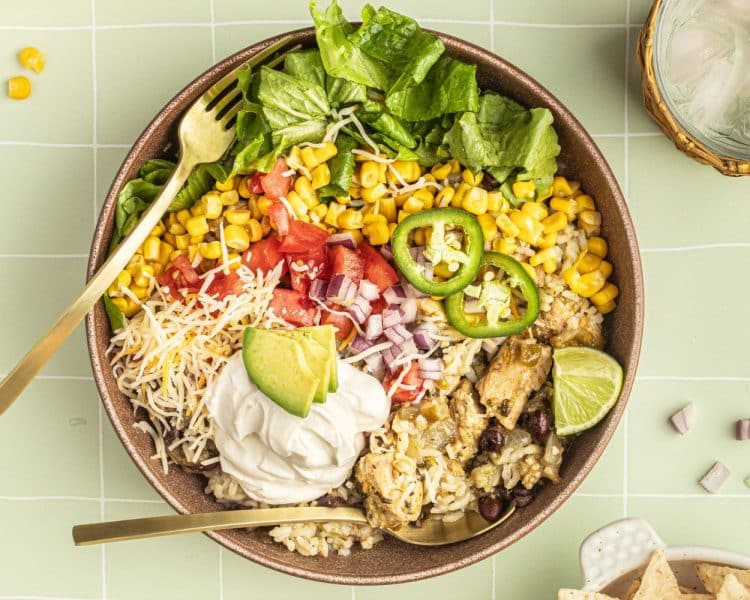 A bowl with a gold fork and spoon and white rice, chicken, black beans, cheese, corn, lettuce, tomatoes, diced onions, sour cream, lime wedges, and sliced jalapeno.