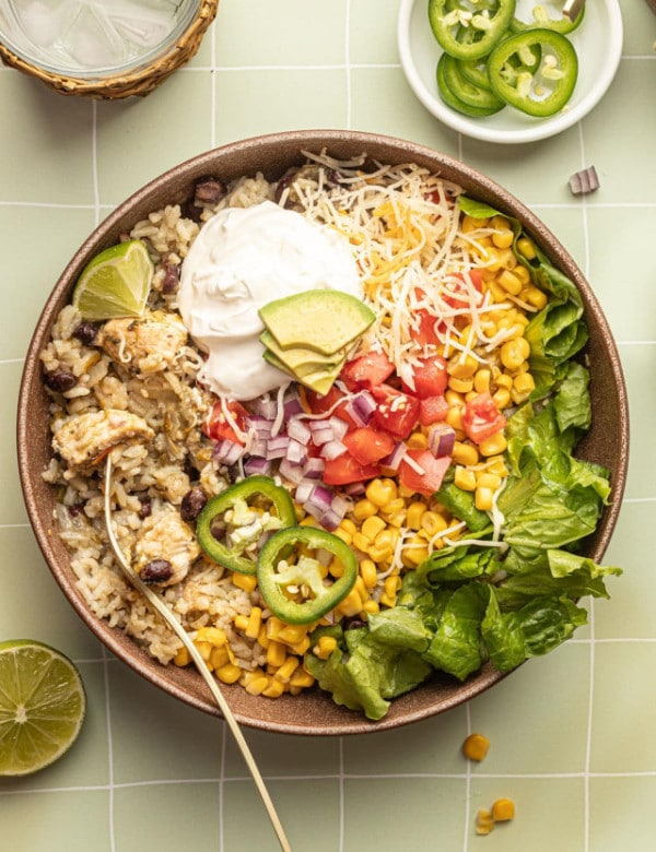 A bowl with a gold fork and spoon and white rice, chicken, black beans, cheese, corn, lettuce, tomatoes, diced onions, sour cream, lime wedges, and sliced jalapeno.