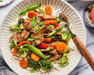 A plated serving of beef and veggie stir fry over white rice in a shallow bowl, garnished with sliced green onions and served with a fork.