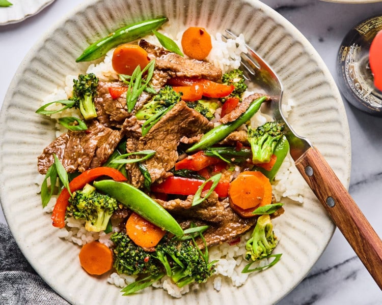 A plated serving of beef and veggie stir fry over white rice in a shallow bowl, garnished with sliced green onions and served with a fork.