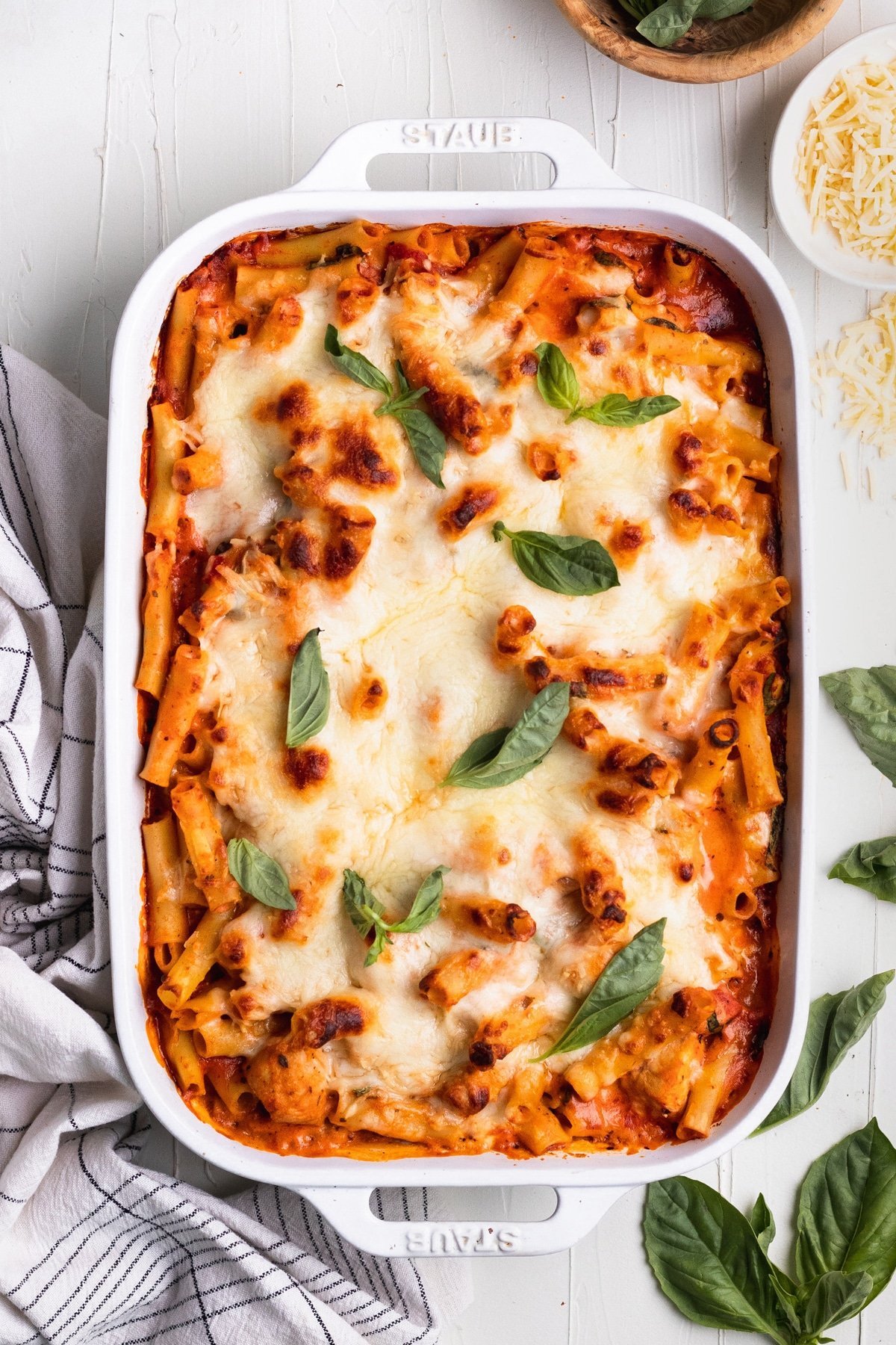 baked ziti in a white casserole dish topped with cheese and fresh basil