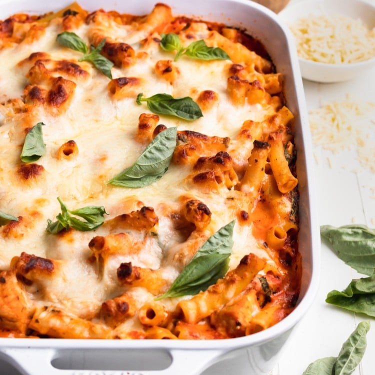baked ziti in a white casserole dish topped with cheese and fresh basil
