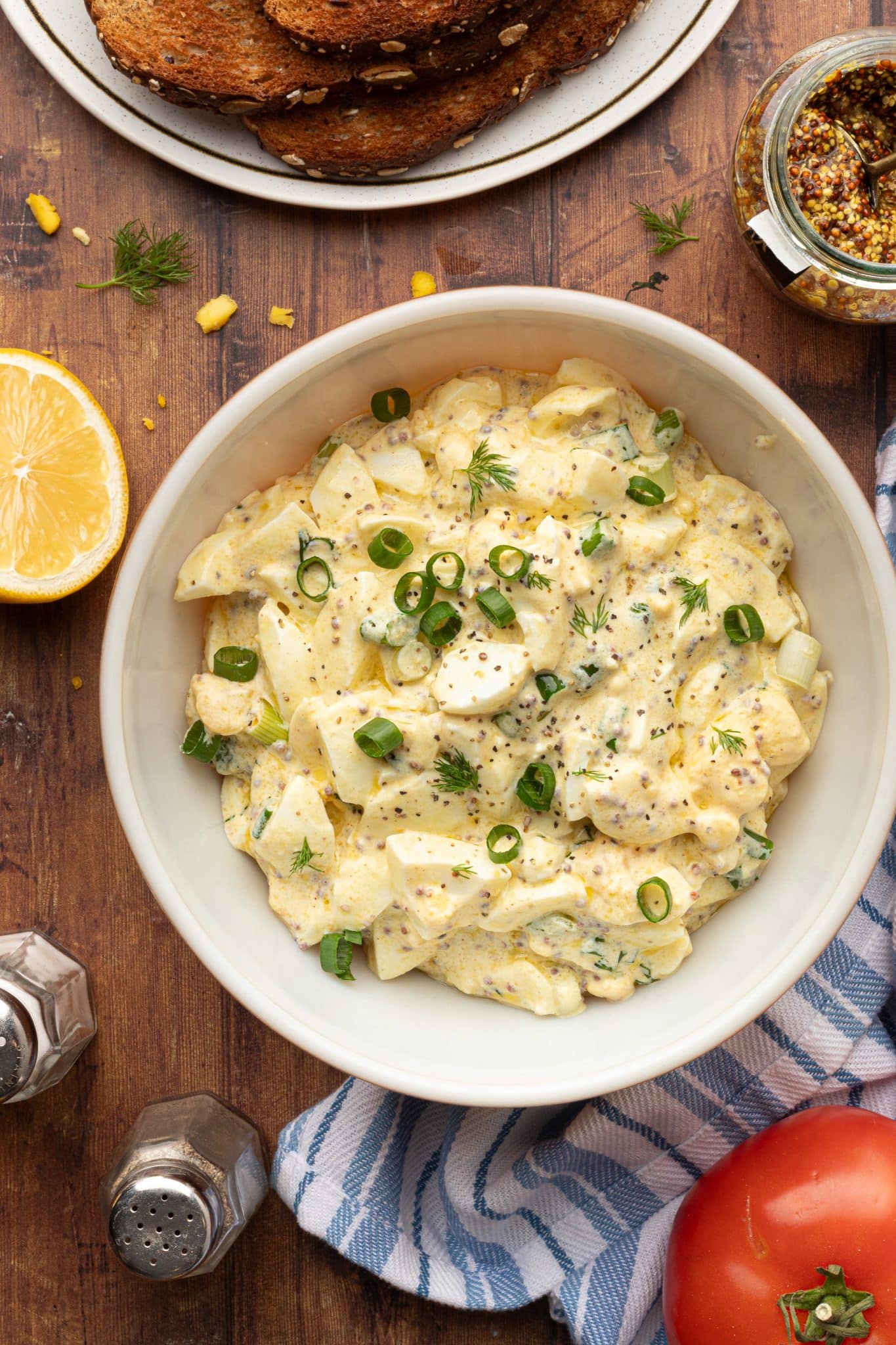 A white bowl filled with a creamy, chopped egg salad garnished with fresh dill and sliced green onions, surrounded by toasted bread, a jar of whole-grain mustard, a halved lemon, salt and pepper shakers, and a ripe tomato on a wooden table.