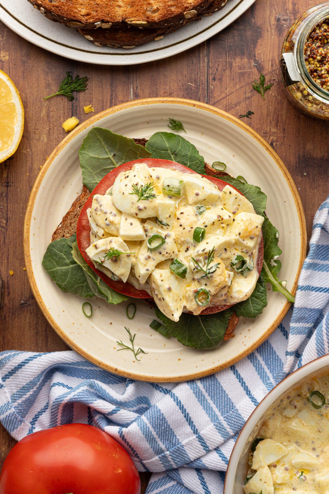 An open-faced egg salad sandwich on toasted bread, topped with tomato slices, leafy greens, and a creamy egg mixture, garnished with fresh dill and green onions. A jar of whole-grain mustard, a lemon half, and a ripe tomato rest nearby on a rustic wooden surface.


