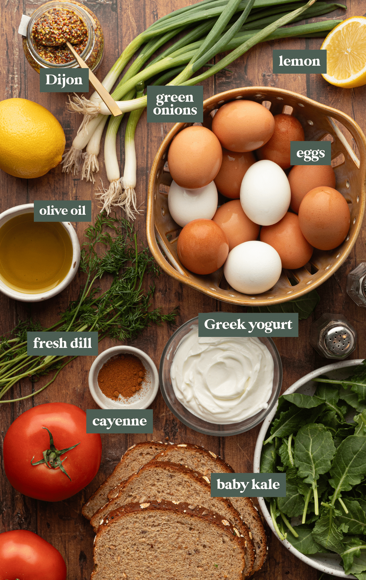 An overhead view of a wooden table displaying labeled ingredients for making egg salad: a bowl of brown and white eggs, Greek yogurt, fresh dill, green onions, tomatoes, baby kale, whole-grain bread slices, a halved lemon, olive oil, Dijon mustard, and a small bowl of cayenne pepper.