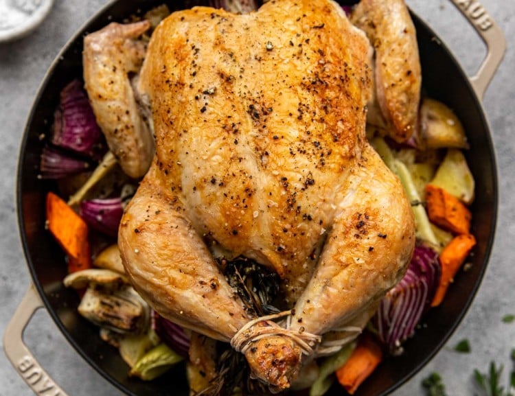 full roasted chicken in a dutch oven surrounded by roasted vegetables