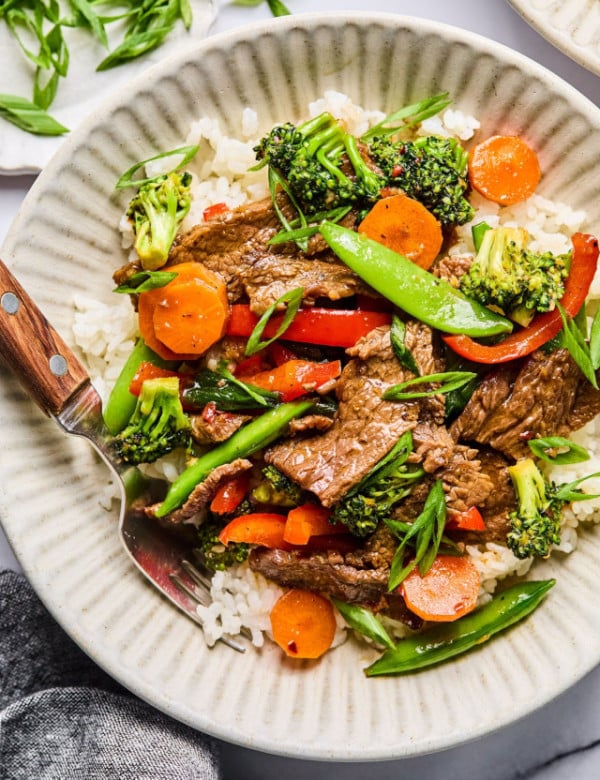 Close-up overhead of a single bowl filled with beef stir fry and rice, showing colorful veggies, beef, and a sprinkle of green onion slices.