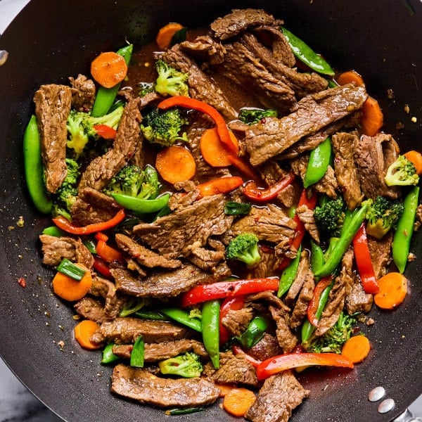Overhead view of a wok filled with beef stir fry featuring seared beef strips, bright carrots, broccoli florets, snap peas, and red bell peppers in a glossy sauce.