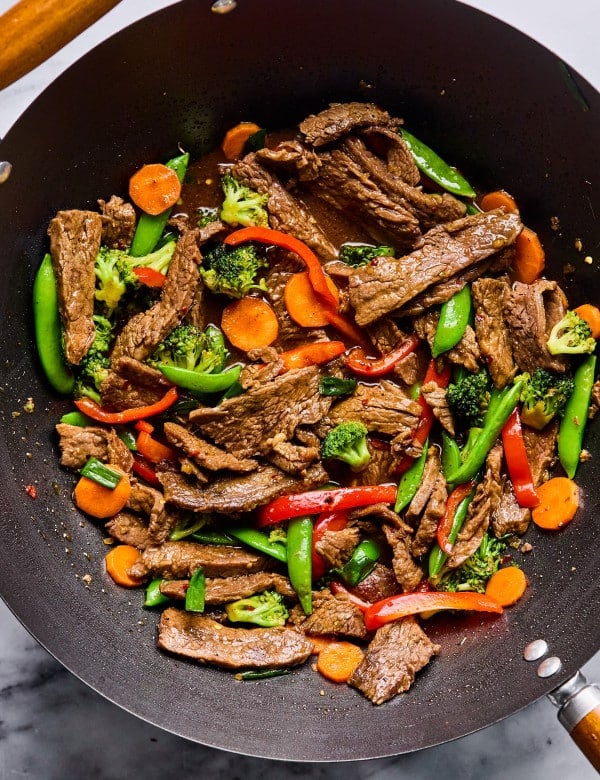Overhead view of a wok filled with beef stir fry featuring seared beef strips, bright carrots, broccoli florets, snap peas, and red bell peppers in a glossy sauce.