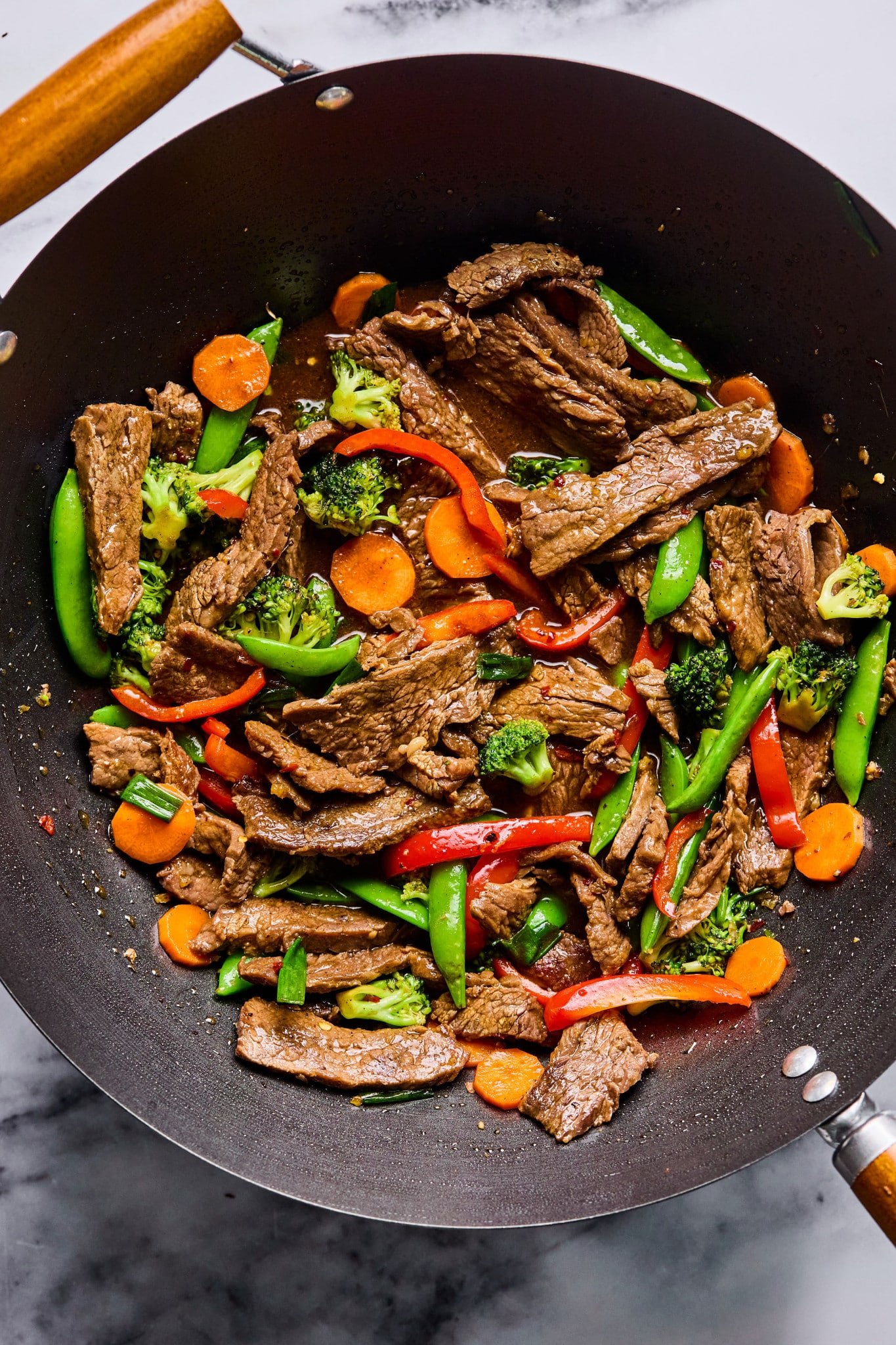 Overhead view of a wok filled with beef stir fry featuring seared beef strips, bright carrots, broccoli florets, snap peas, and red bell peppers in a glossy sauce.