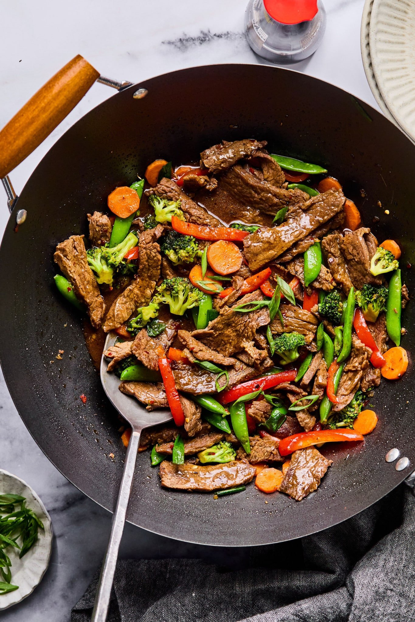 Overhead view of the beef stir fry in a wok with a metal serving spoon, ready for plating, surrounded by a soy sauce bottle and plate of sliced green onions.