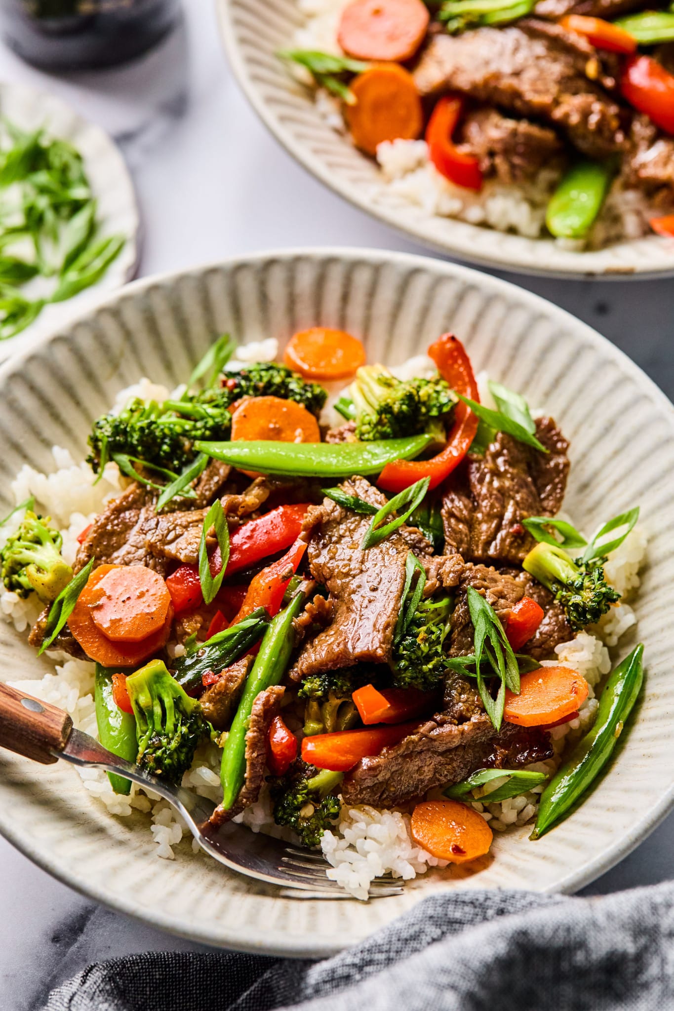 Side angle of a bowl of beef stir fry over rice, focusing on the tender beef, carrots, broccoli, and snap peas, with a fork resting in the bowl.