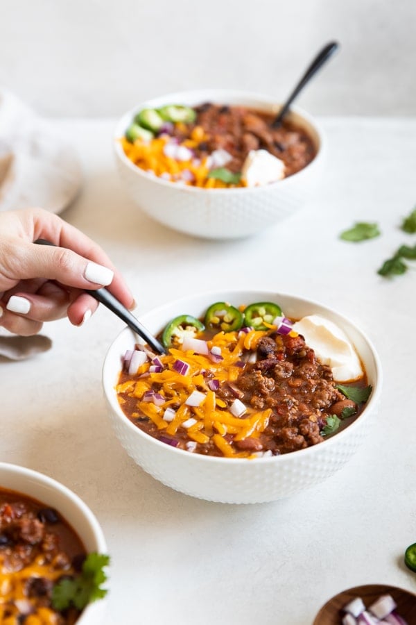 4 bean spicy crockpot chili in white bowls topped with cheese, onions and sour cream 