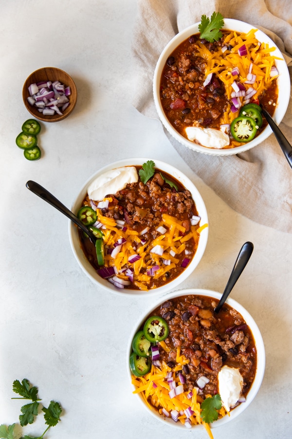 4 bean spicy crockpot chili in white bowls topped with cheese, onions and sour cream 