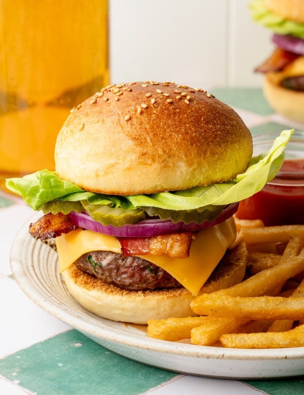 Homemade cheeseburger on a plate with butter lettuce, pickles, red onion, bacon, and cheddar cheese, with a side of fries and a glass of beer in the background.