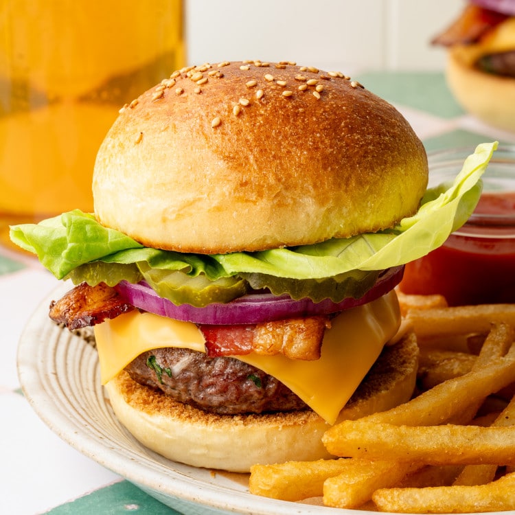 Homemade cheeseburger on a plate with butter lettuce, pickles, red onion, bacon, and cheddar cheese, with a side of fries and a glass of beer in the background.