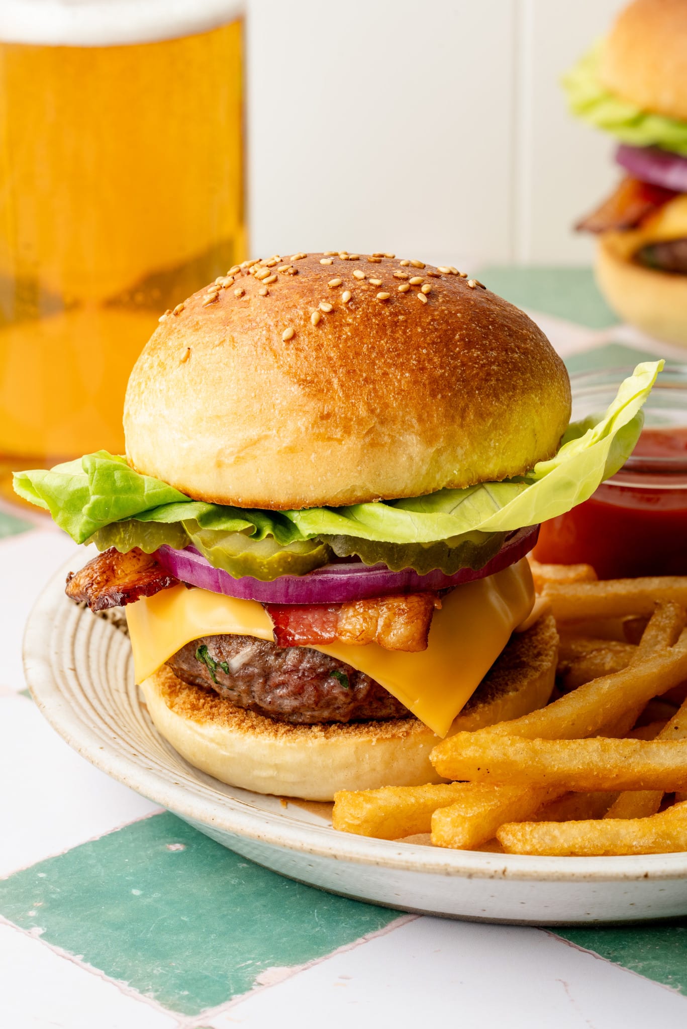 Homemade cheeseburger on a plate with butter lettuce, pickles, red onion, bacon, and cheddar cheese, with a side of fries and a glass of beer in the background.
