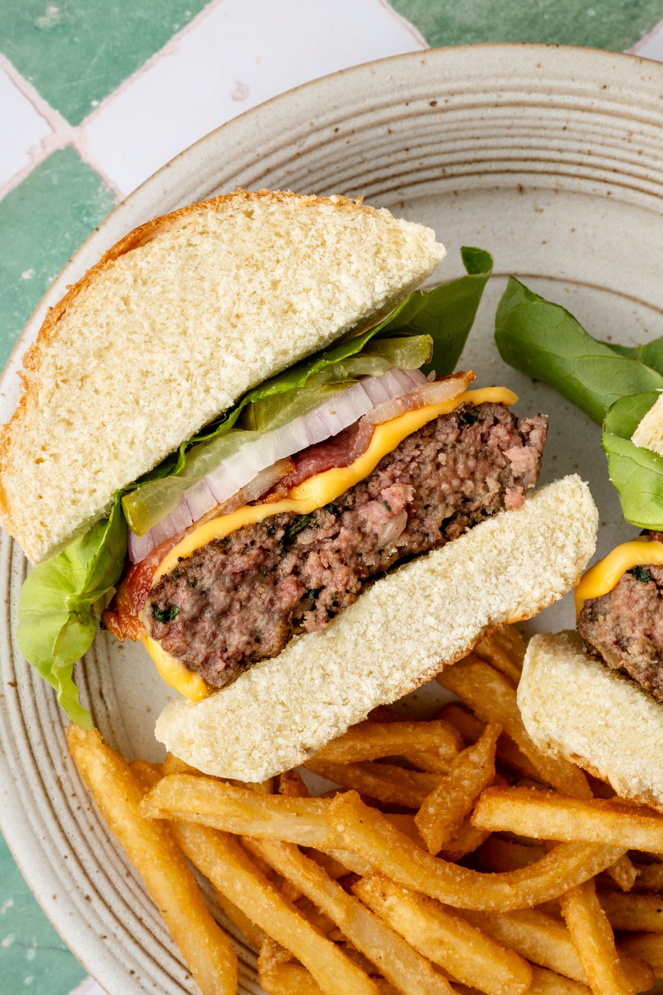 Sliced homemade cheeseburger on a plate showing a juicy beef patty layered with cheddar cheese, bacon, pickles, red onion, and lettuce, served with a side of fries.