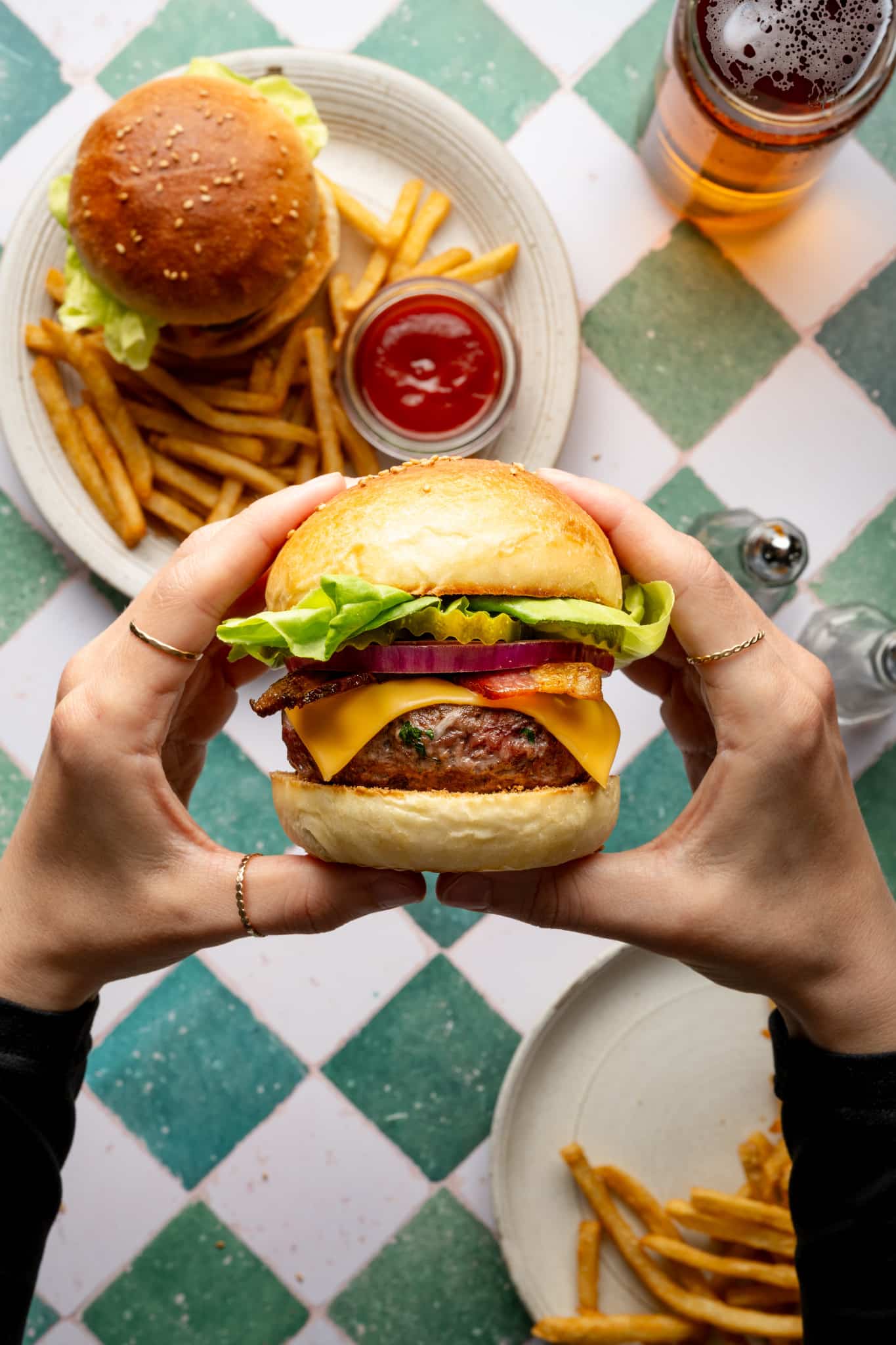 Hands holding a homemade cheeseburger filled with lettuce, pickles, red onion, bacon, and cheddar cheese. A second plate with fries and ketchup is seen below.