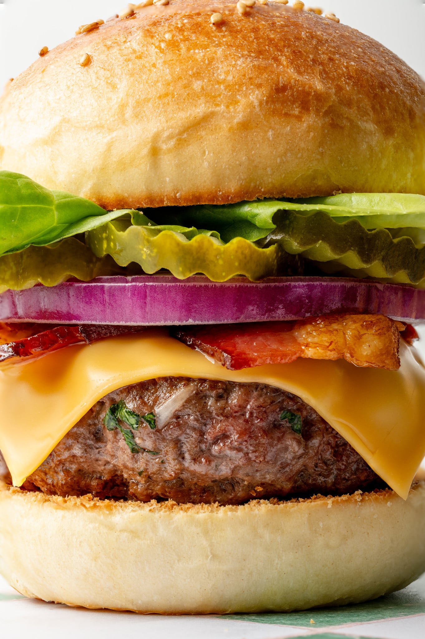 Extreme close-up of a juicy homemade cheeseburger showing layers of butter lettuce, pickles, red onion, crispy bacon, melted cheese, and a thick beef patty on a toasted bun.