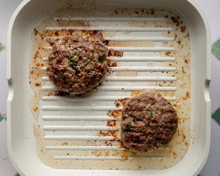 Two beef patties cooking on a white grill pan with seared grill marks and browning visible, showing the burgers partway through cooking.