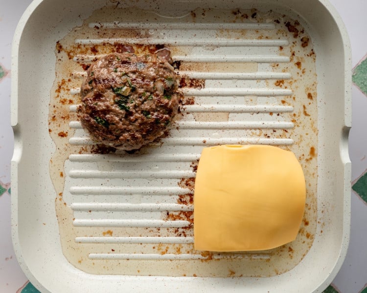 One cooked beef patty on a grill pan and another topped with a slice of cheddar cheese beginning to melt, showing the cheese-melting step of the burger process.