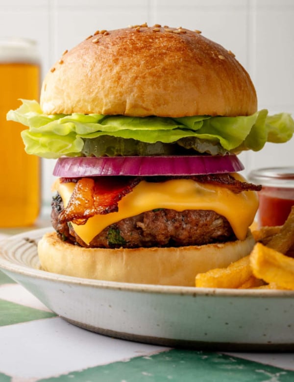 Side view of a tall homemade cheeseburger stacked with butter lettuce, pickles, red onion, crispy bacon, and melted cheddar cheese, with fries and beer blurred in the background.