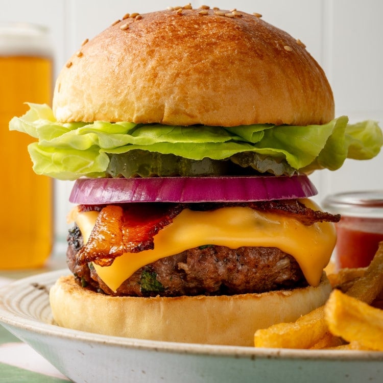 Side view of a tall homemade cheeseburger stacked with butter lettuce, pickles, red onion, crispy bacon, and melted cheddar cheese, with fries and beer blurred in the background.