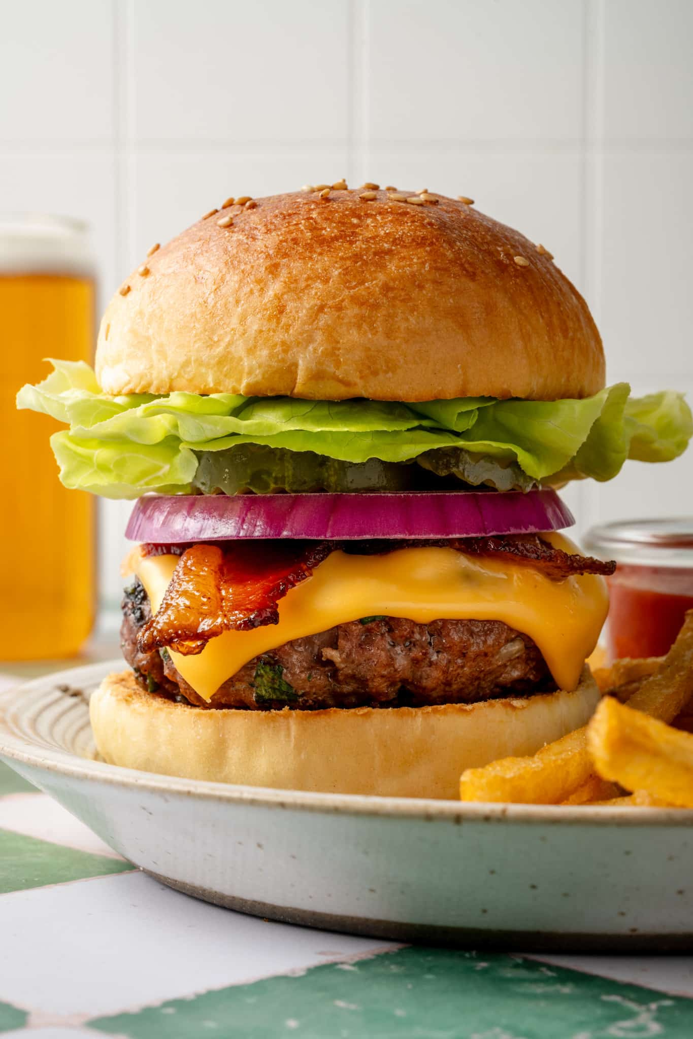 Side view of a tall homemade cheeseburger stacked with butter lettuce, pickles, red onion, crispy bacon, and melted cheddar cheese, with fries and beer blurred in the background.