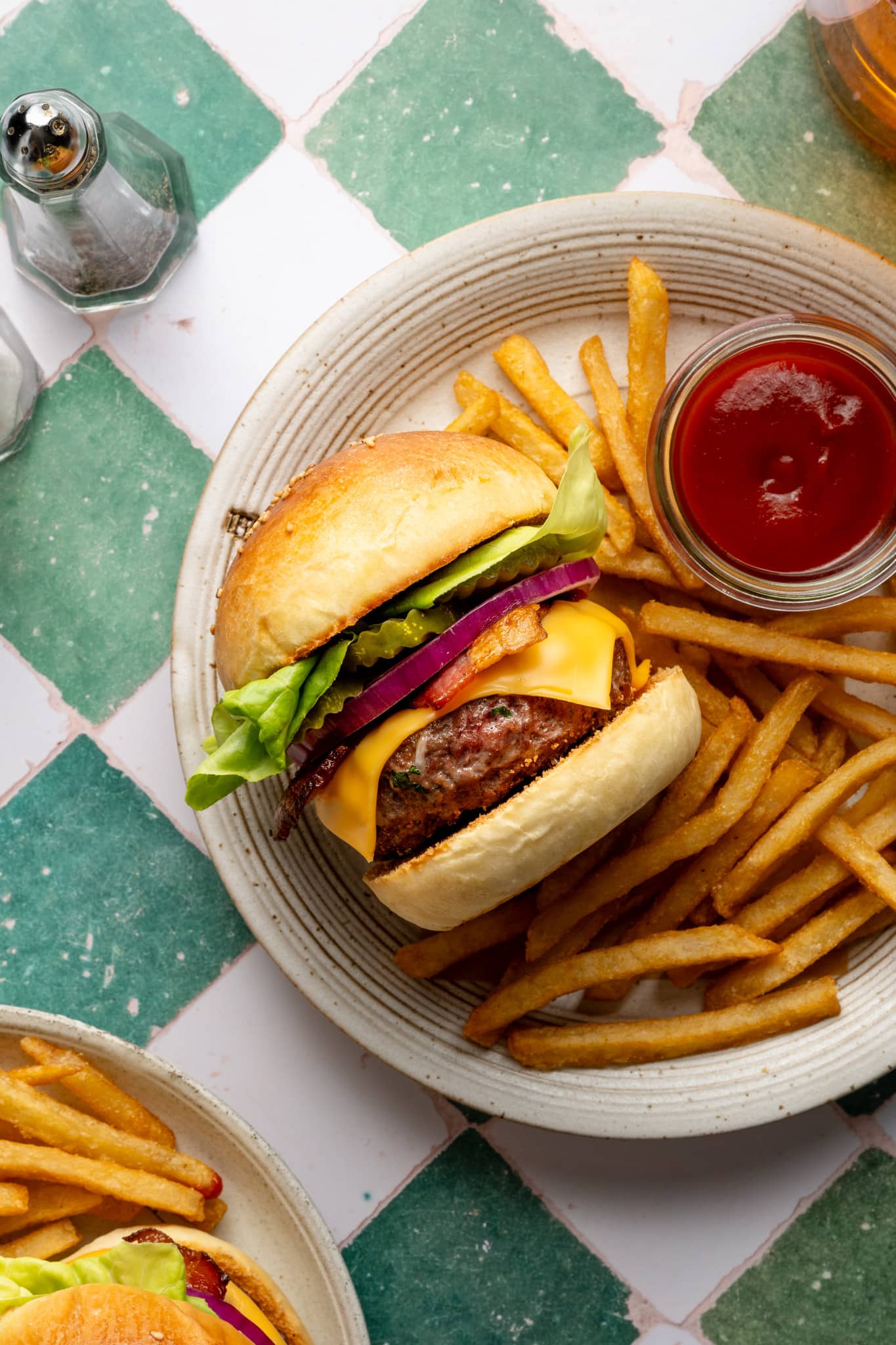 Top-down view of a plate with a homemade cheeseburger, crispy fries, and a small jar of ketchup. The burger features butter lettuce, onion, bacon, pickles, and melted cheese.