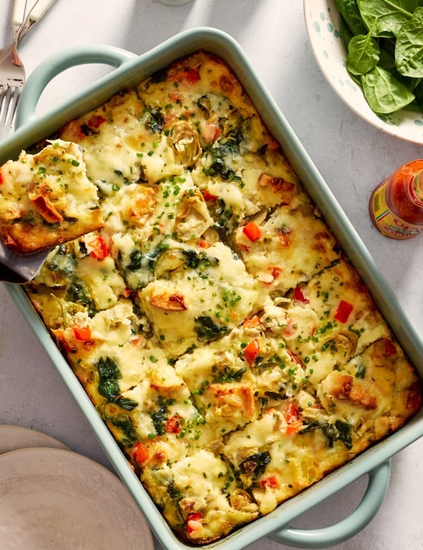 Overhead photo of a baked overnight artichoke spinach egg bake in a light blue casserole dish with a square slice lifted on a spatula. The cheesy breakfast casserole is filled with spinach, marinated artichokes, red bell peppers, toasted English muffin pieces, and chopped chives sprinkled on top.