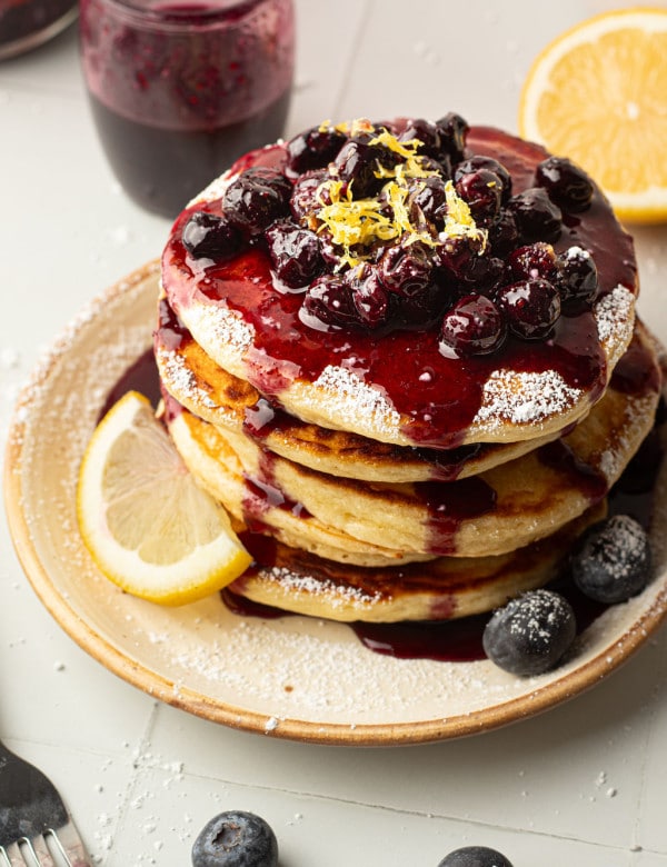 A top-down view of a pancake stack covered in blueberry compote, powdered sugar, and lemon zest. The plate is garnished with fresh blueberries and a lemon wedge. Blueberry sauce has pooled at the base of the pancakes.