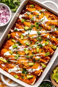 white casserole dish filled with red enchiladas topped with a drizzle of sour cream, chopped avocado, and cilantro