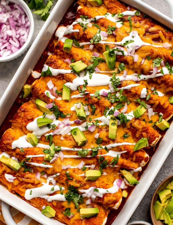 white casserole dish filled with red enchiladas topped with a drizzle of sour cream, chopped avocado, and cilantro