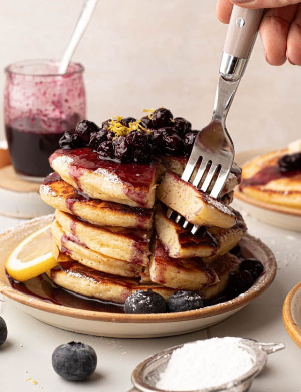 A hand cutting into a stack of pancakes, revealing the soft, fluffy interior. The pancakes are covered in blueberry compote, lemon zest, and powdered sugar. A jar of blueberry sauce and a second plate of pancakes are in the background.