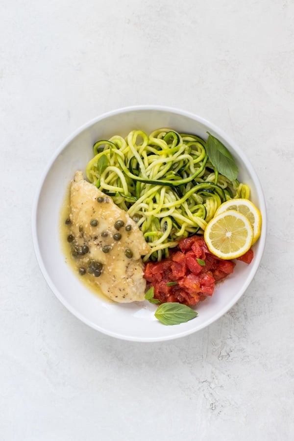 chicken piccata in a white bowl with diced tomatoes and zoodles