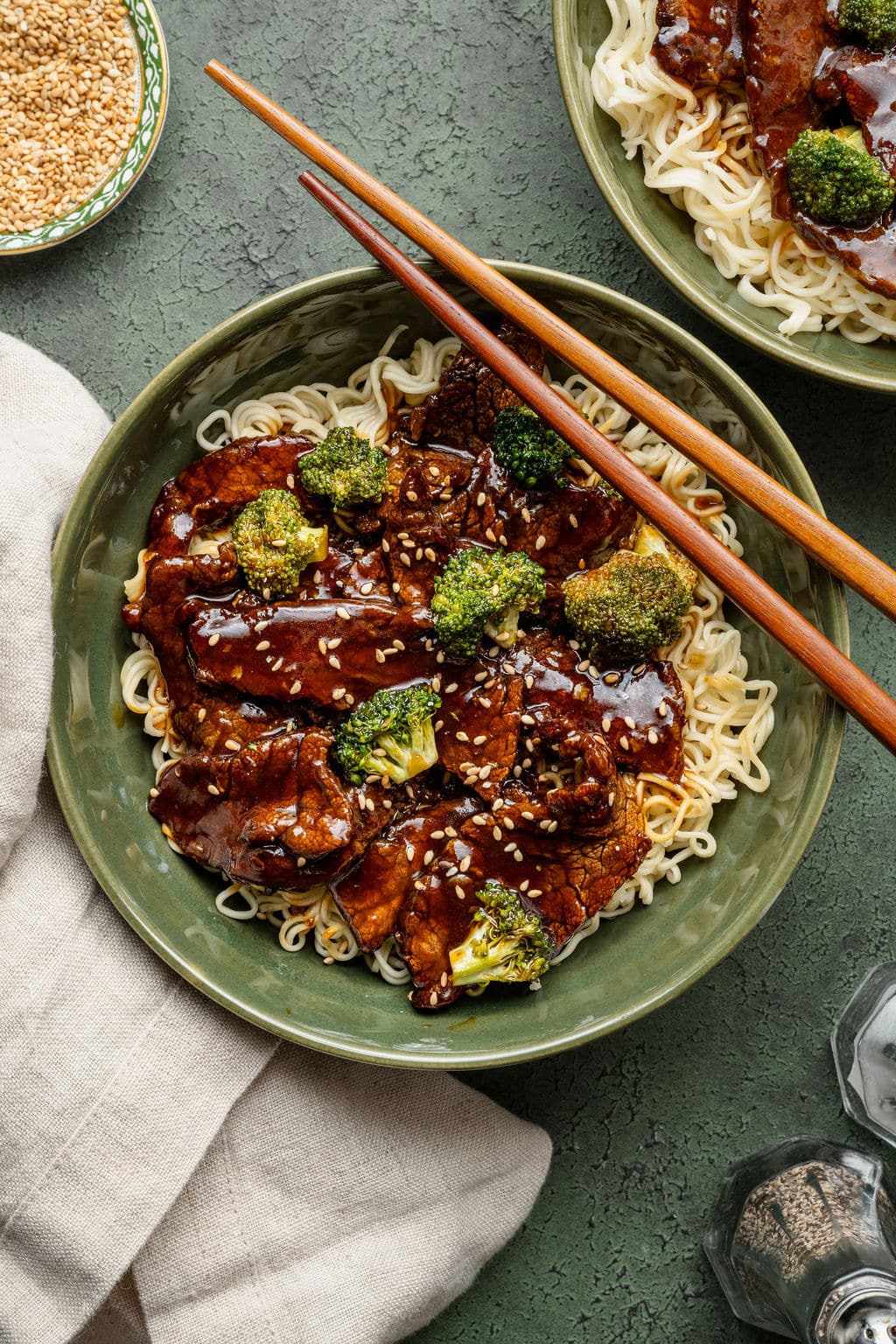 Bowl of beef and broccoli over ramen noodles, garnished with sesame seeds and served with chopsticks resting on the rim.