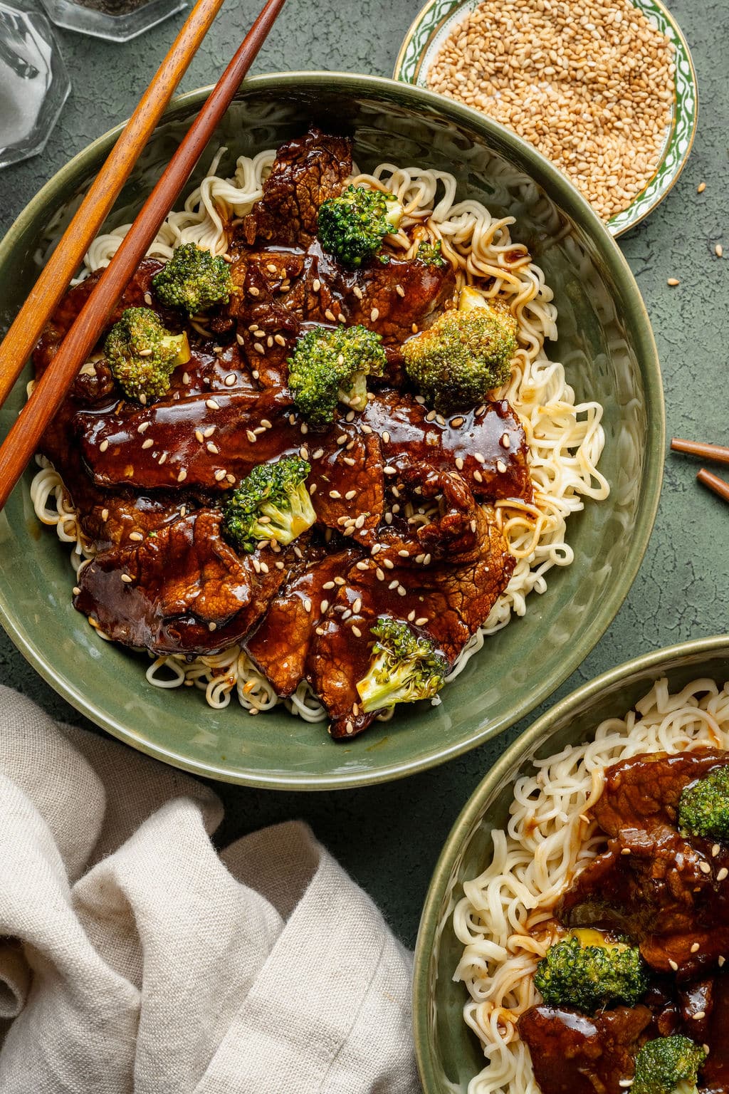 Top-down view of two green bowls filled with beef and broccoli over noodles, sprinkled with sesame seeds, surrounded by small dishes and napkins.