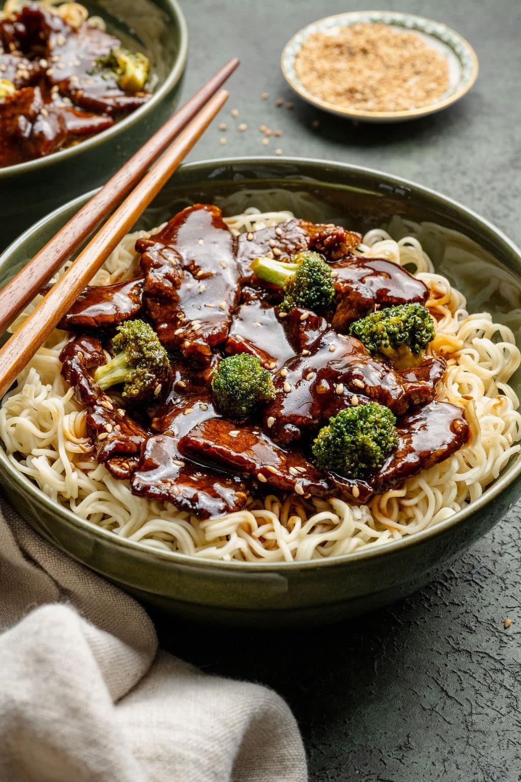 Side view of beef and broccoli noodles in a green bowl with chopsticks, showing glossy sauce and sesame seed garnish.