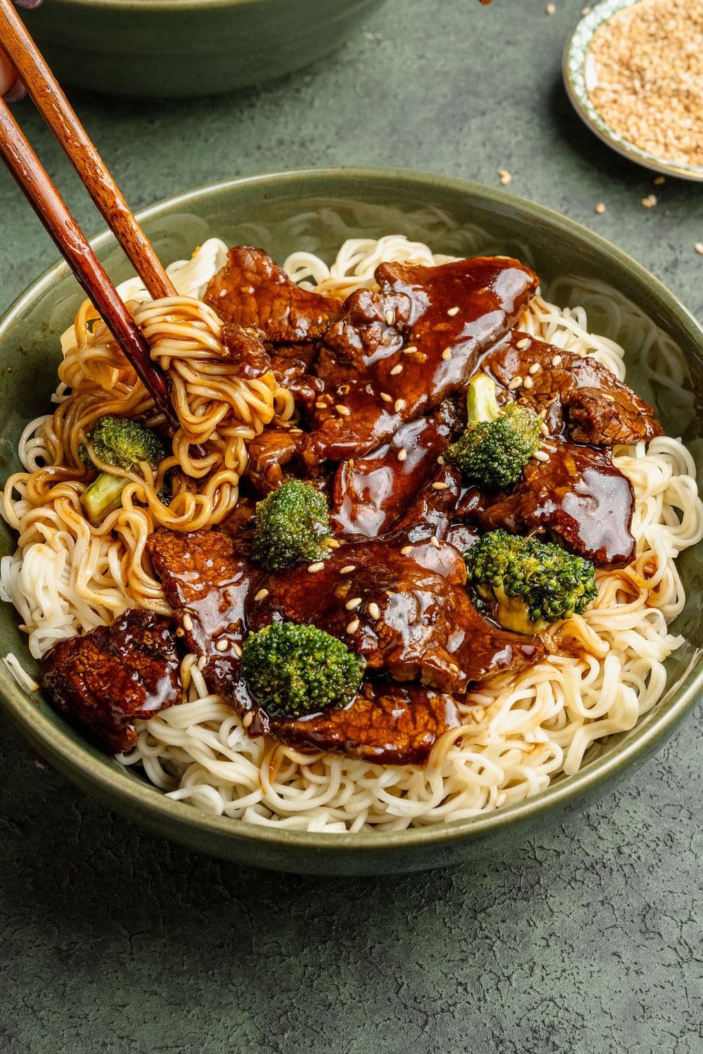 Close-up of beef and broccoli noodles with chopsticks lifting a bite of noodles coated in savory sauce.