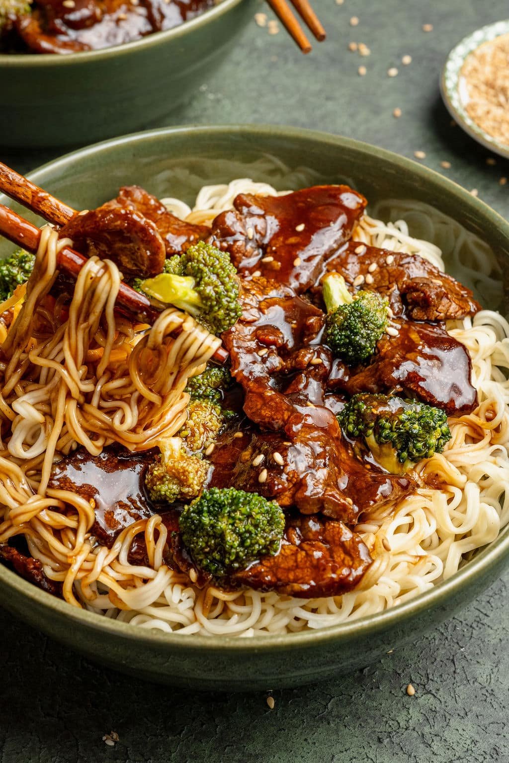 Tight shot of chopsticks lifting noodles from a bowl of beef and broccoli, highlighting the glossy sauce, sesame seeds, and tender pieces of beef.