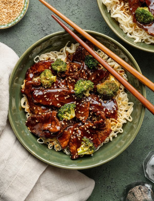 Bowl of beef and broccoli over ramen noodles, garnished with sesame seeds and served with chopsticks resting on the rim.