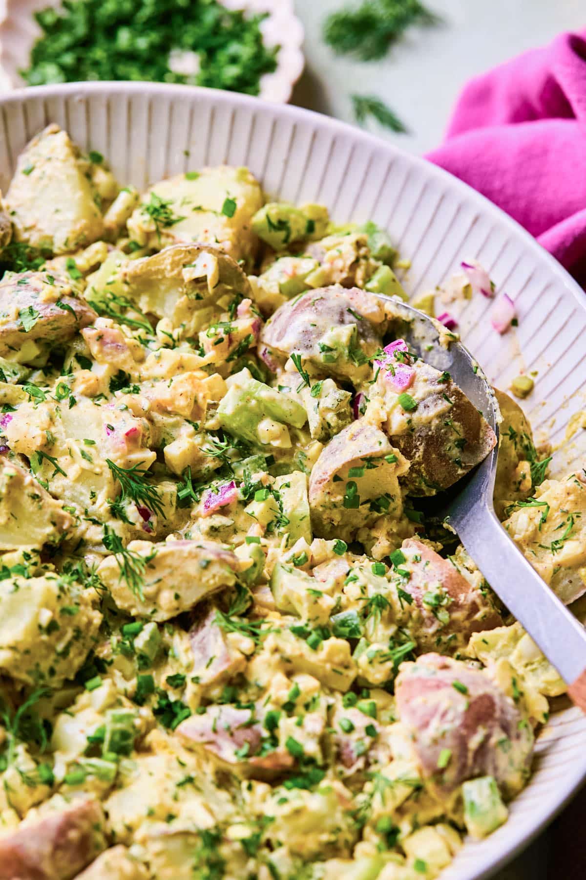 Spoon in a bowl of potato salad made with red potatoes, chopped celery, pickles, red onions, and fresh herbs.