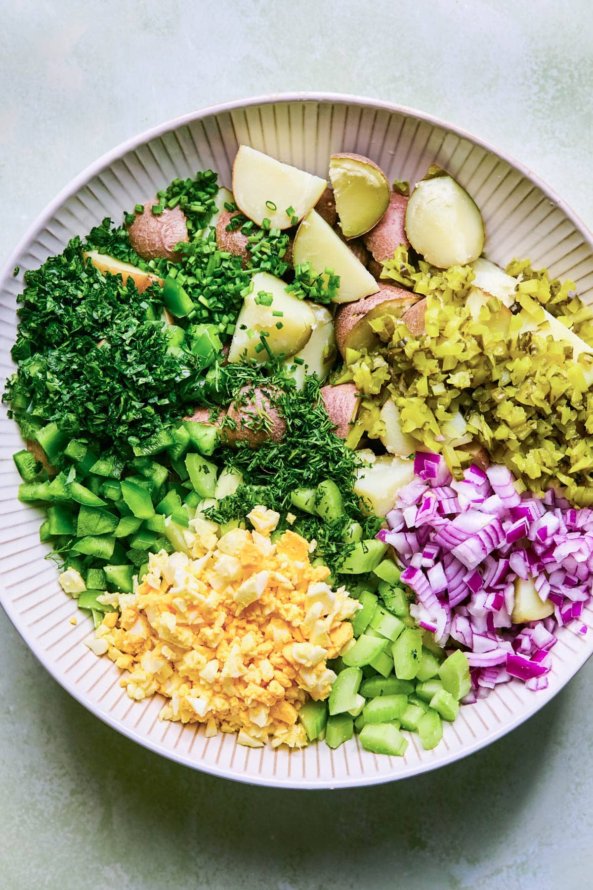 Ingredients including chopped parsley, chives, red potatoes, pickles, red onion, hard boiled eggs and celery in a large bowl.