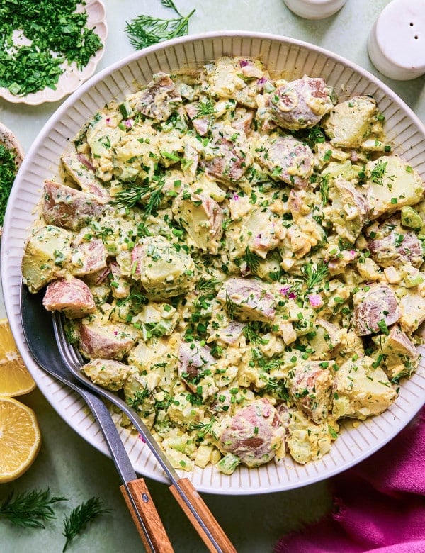 White bowl full of creamy, herby potato salad with serving utensils at the side.